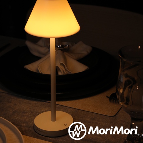 白色圓形底座的LED傘型造型燈，燈罩為淺黃色，散發溫暖光線，置於餐桌上，背景為黑色餐盤和白色餐巾。