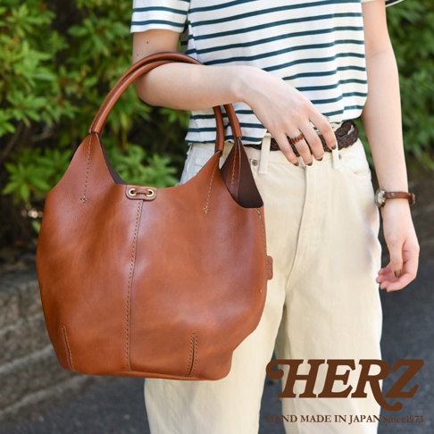 職人手作皮革球形肩掛托特包，顏色為棕褐色。包款採用皮革材質，具有圓潤的造型和手工縫線細節，搭配品牌標誌。