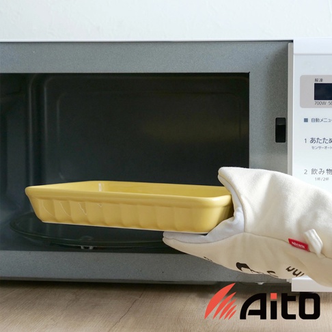 一隻戴著白色隔熱手套的手，正將一個黃色釉彩的美濃燒萬用長方烤盤放入微波爐中。烤盤邊緣有垂直凹槽設計。