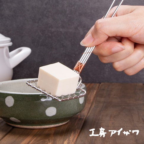 一只手正用不鏽鋼豆腐杓撈起一塊白色豆腐，豆腐杓網格為方形，手柄處以銅線纏繞裝飾。背景為綠色有白色圓點的陶瓷碗和木質桌面。