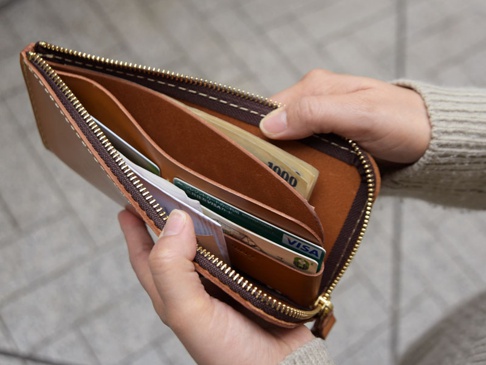 雙手拿著一個棕色皮革L型拉鍊長夾，夾內有鈔票和信用卡，拉鍊為金色，縫線為白色。