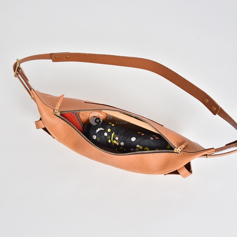 一個棕褐色的皮革斜背包，拉鍊打開，裡面放著一個印有彩色圓點的摺疊傘和一些其他小物。