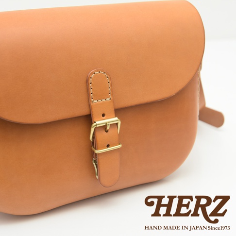 職人手作皮革經典馬鞍包，淺棕色，圓潤飽滿的包身，搭配黃銅釦環與皮質錶帶，簡潔的縫線展現細緻工藝。