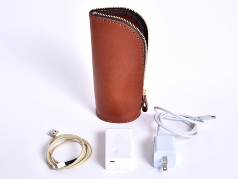 職人手作皮革直立式收納筆袋，經典棕色，拉鍊開合。旁邊放置一個白色方塊充電器、一個白色充電線和一個米色充電線。