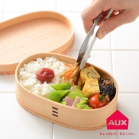 一只手正用金屬夾子夾取木製便當盒中的燉菜，便當盒內有米飯、醃梅、煎蛋捲、青豆、火腿和番茄。