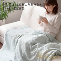 一位女性坐在白色沙發上，身上蓋著一條淺藍色、輕盈的四層紗毛巾毯。毛巾毯呈現柔軟皺褶的紋理。