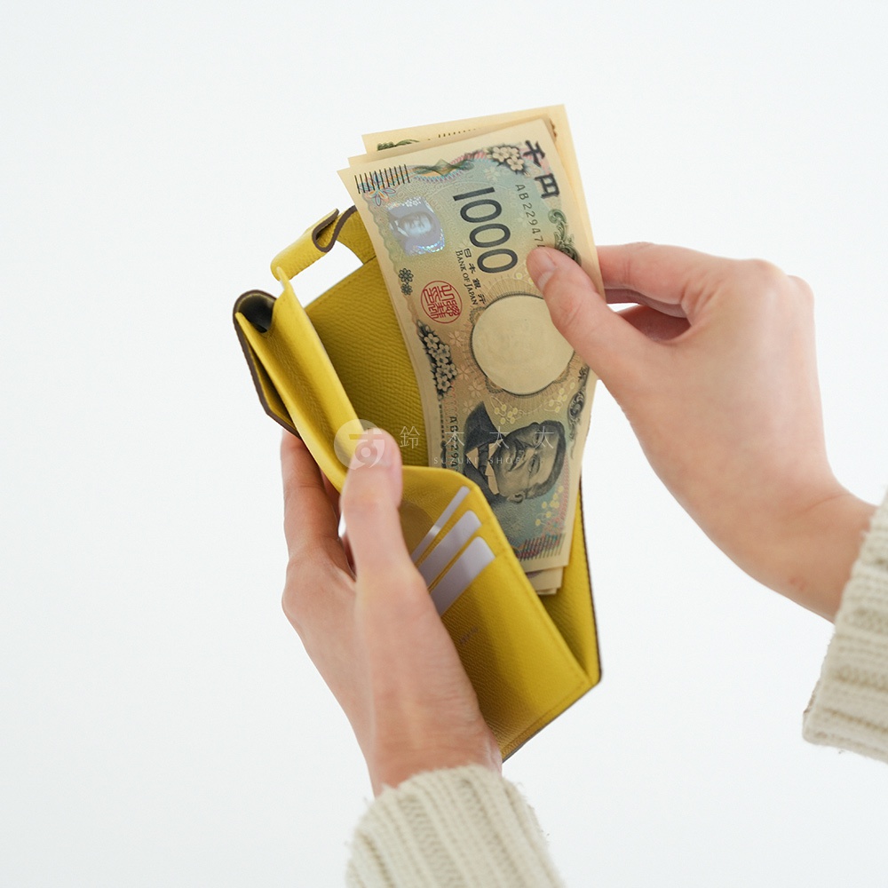 黃色皮革短夾中，一只手正在放入兩張日幣千元鈔票，鈔票上有著千圓的數字標示。