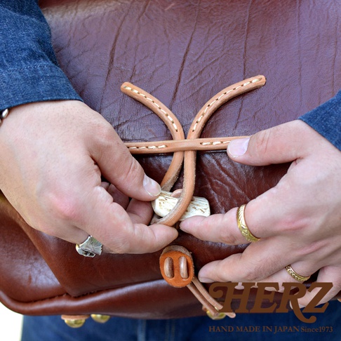 職人手作皮革鹿角扣繩郵差包，深棕色皮革包身，束繩以鹿角狀的扣飾固定，手工縫線清晰可見，展現精緻工藝。