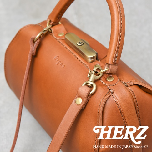 職人手作皮革Organ兩用口金手提包，採用光滑的棕色皮革，配有金色金屬口金鎖扣、提把和可拆卸肩帶。包身呈現圓筒狀，皮革縫線清晰可見。