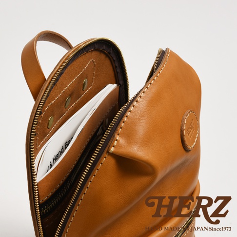 職人手作皮革大開口圓角後背包，展示著溫潤的淺棕色皮革，拉鍊部分為黃銅色。包包內部有隔層，可放置卡片或小冊子。