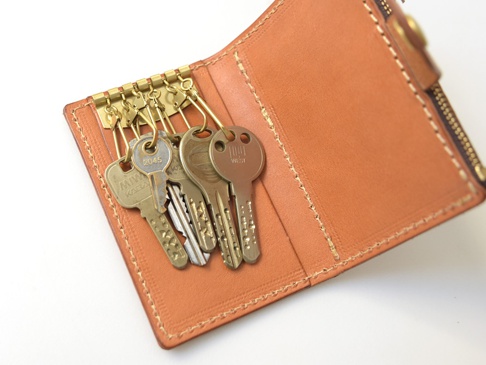 職人手作的淺棕色皮革鑰匙包，內有六個黃銅鑰匙鉤，鉤上掛有數把金屬鑰匙，皮革表面有縫線及拉鍊細節。