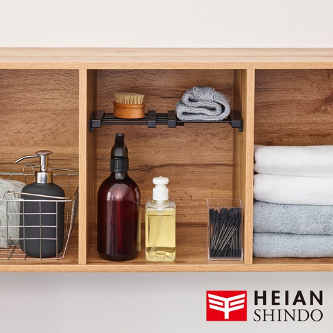 木紋層架上展示著一個黑色按壓瓶皂液器，置於銀色金屬置物籃內；一個深棕色噴霧瓶及一個透明方形瓶裝黃色液體，旁為裝有黑色棉花的透明盒；最右側疊放著白色及淺藍色毛巾。