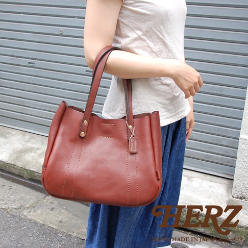 職人手作皮革仕女托特包，棕色，由皮革製成，具有雙提把和縫線細節，搭配女性穿著。