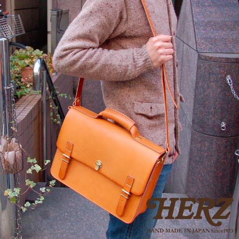 駝色皮革手提/斜背包，正面有兩個皮帶扣和一個鎖扣，搭配同色系皮革提把和可拆卸斜背帶。