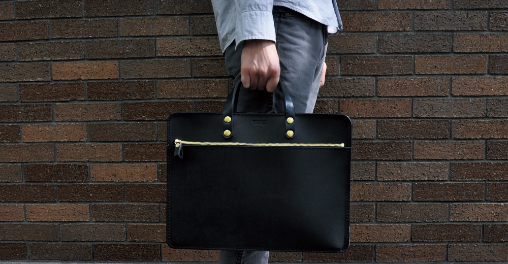 黑色皮革兩用商務公事包，配有金色拉鍊和黃銅鉚釘，可手提或肩背，適合商務場合。