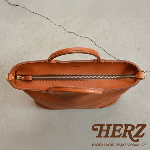 日本HERZ手工皮革Organ兩用拉鍊托特包，採用棕色皮革製成，頂部有拉鍊開口，兩側設有提把，置於灰色水泥地面上。