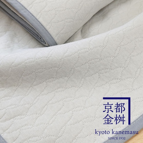 象牙白色天絲棉絎縫單人毯，表面有著流線型的壓紋圖案，邊緣為淺灰色滾邊。右下角印有藍色框線的金色日文「京都金桝」及英文「kyoto kanemasu」字樣，並標示「SINCE 1932」。