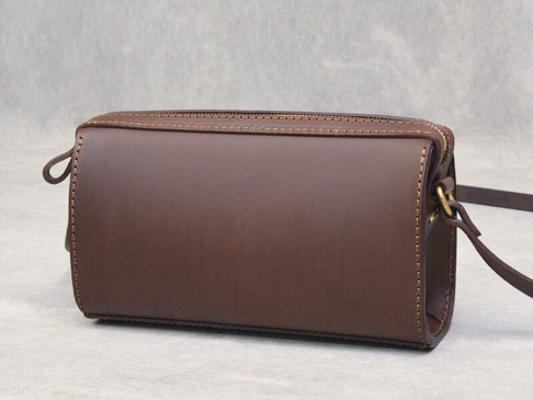 咖啡色皮革製成的拉鍊單肩包，包身呈現圓筒狀，側面有縫線裝飾，附有皮革背帶與金屬掛環。