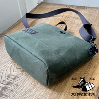 一個綠色的純綿帆布迷你側背包，帶有深灰色織帶和黑色金屬扣件，包包正面有一個品牌標籤。