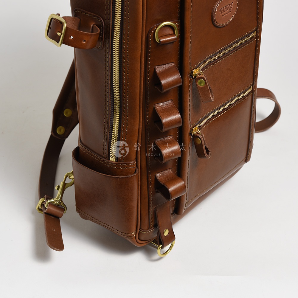 職人手作的棕色皮革兩用後背包，有著黃銅色拉鍊、釦環與 D 型環，側面有四個皮質環扣，右側有兩個拉鍊口袋，左側有一個開放式口袋，可見品牌標誌。
