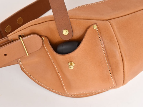 職人手作皮革斜背包，呈現天然原色，材質為真皮。包款設有外側小口袋，可放入耳機等小物，並以銅質釦件與縫線裝飾，展現手工質感。