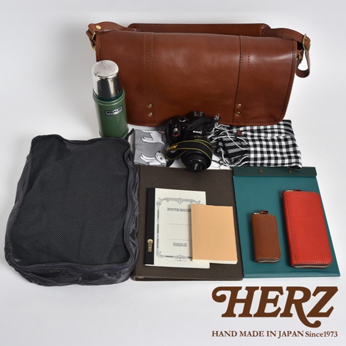 職人手作皮革復古郵差包，搭配綠色保溫瓶、黑色網狀收納包、相機、格紋頭巾、筆記本、現金夾和鑰匙包。
