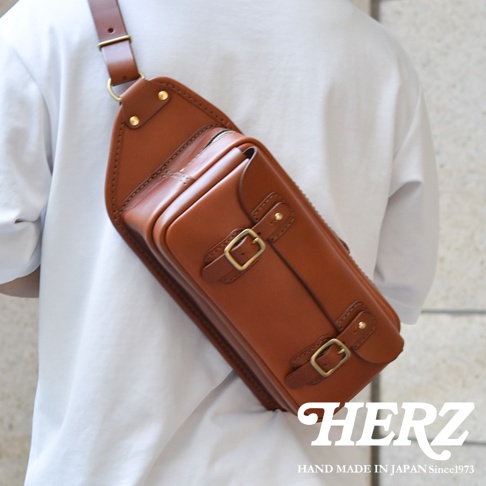 職人手作皮革騎士隨身斜背包，採用棕色皮革製成，設有拉鍊主袋和兩個帶有黃銅扣的翻蓋前袋，肩帶可調節。