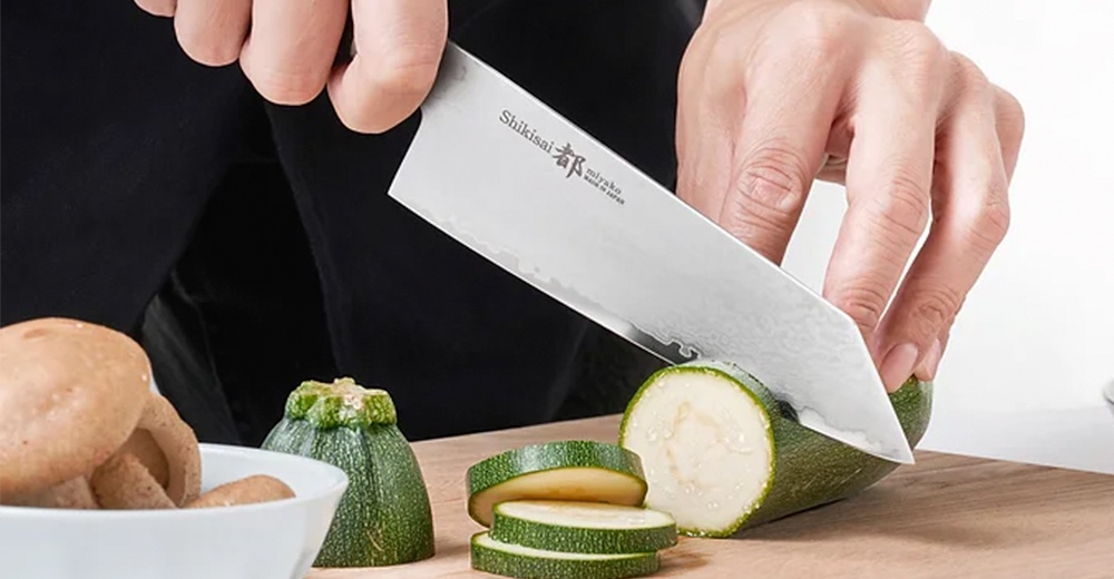 手持一把銀色金屬材質的 Bunka 文化刀，正在切片綠色櫛瓜。刀刃上印有「Shikisai 都 miyako made in JAPAN」字樣。一旁有裝有香菇的白色碗。