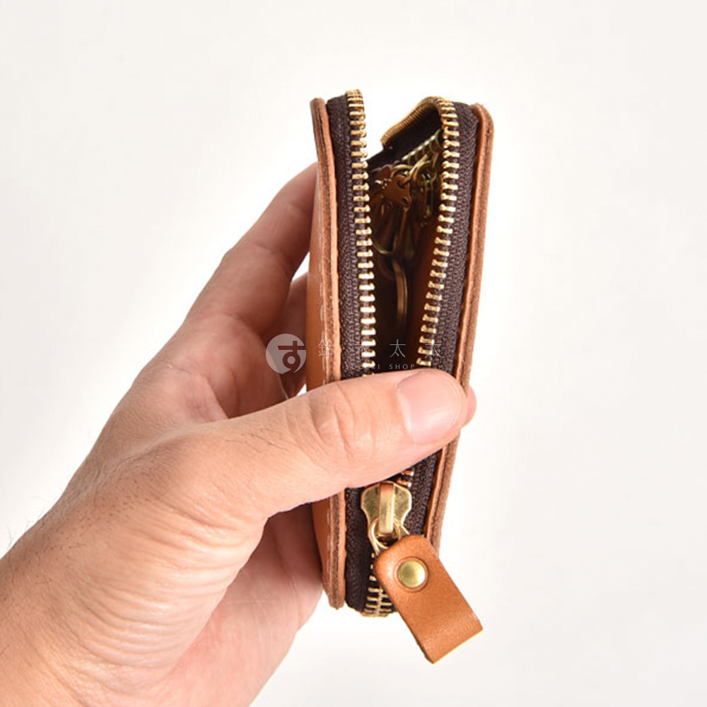 一手拿著棕色皮革鑰匙包，拉鍊半開，露出內部幾個掛著鑰匙的金屬掛鉤。鑰匙包採用黃銅色拉鍊和圓形金屬釘裝飾。