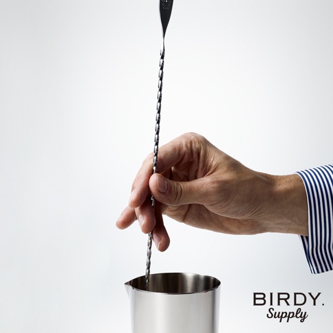 一手持銀色螺旋紋不鏽鋼調酒攪拌匙，匙柄末端為叉狀，置於銀色不鏽鋼量杯中，背景為白色。