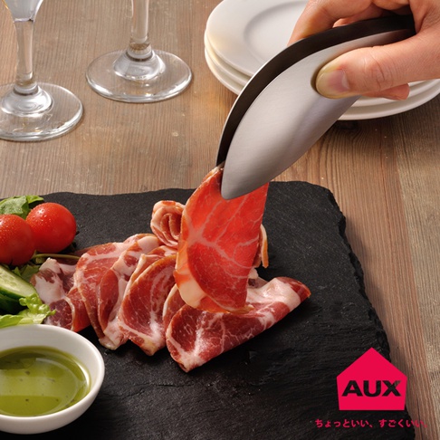 一只手使用不銹鋼材質的leye 不沾桌調理幫手夾，夾起一片薄切的粉紅色生肉，生肉上帶有明顯的白色脂肪紋理，下方是堆疊的生肉片、幾顆小番茄、小黃瓜片、生菜以及一小碗綠色醬汁，所有食材都擺放在黑色石板上。