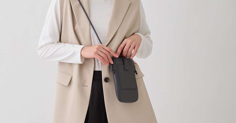 一位女士穿著米色無袖西裝外套，內搭白色長袖上衣，她左手拿著一個黑色皮革手機包，包包上有一個小磁釦和一個壓紋圖案。