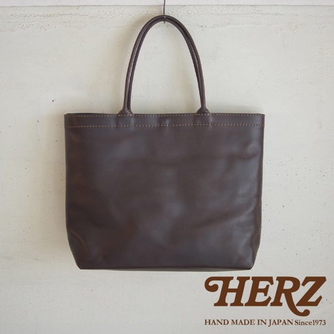 深棕色皮革製成的托特包，正面簡潔無裝飾，上方有雙提把，品牌HERZ標誌印在右下角。
