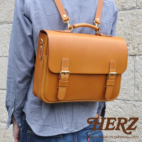 一件淺棕色皮革製成的三用商務公事包，具有極簡風格，配有兩個翻蓋扣帶、一個頂部提把和可拆卸的肩帶。