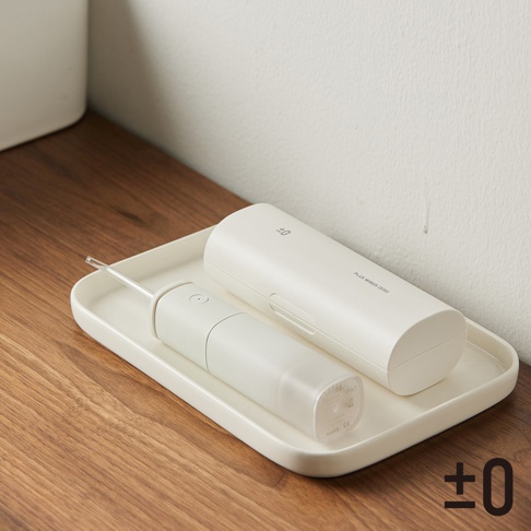 圖片中的白色沖牙機和充電盒擺放在一個淺色托盤上，托盤置於木紋桌面上。沖牙機主體為白色，帶有透明儲水槽，頂部有噴嘴。充電盒也是白色，表面有紋理，上面印有品牌標誌和文字。