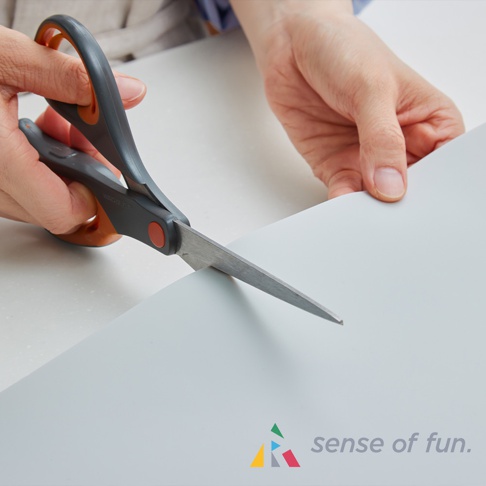 灰色與橘色手柄的剪刀正在裁剪一張淺灰色、光滑表面的長方形保護墊。