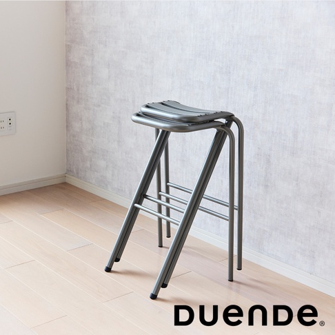 金屬材質的灰色高腳吧檯椅，椅面為弧形設計，椅腳為交叉結構，帶有橫向支撐桿，放在木質地板上，背景為淺色紋理牆面。