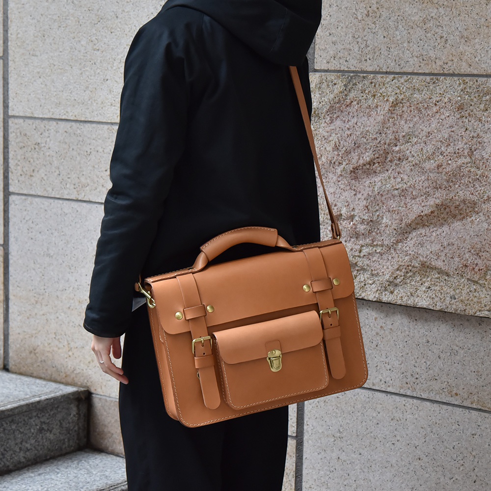 皮革學院風兩用公事包，採用 tan 色皮革製作，配有頂部提把、可拆卸肩背帶，以及前置翻蓋口袋和金屬扣環。