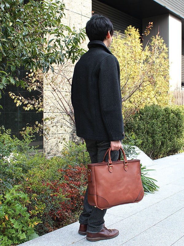 一位男士手提一個棕色皮革托特包，包包有兩個手柄，包包側面有一個可拆卸的肩帶扣環，整體呈現經典商務風格。