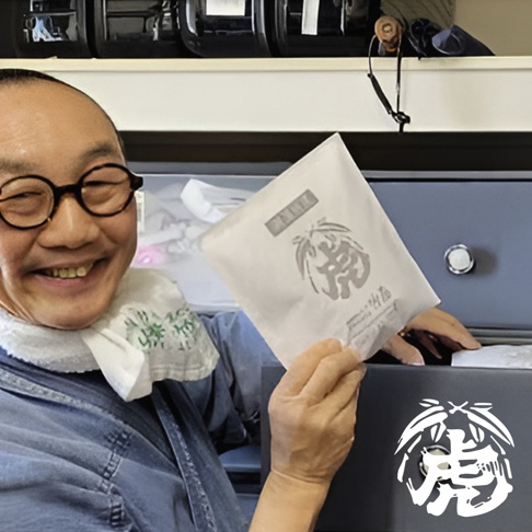 一個男人拿著印有虎形圖案的白色包裝袋，他戴著圓框眼鏡，身穿牛仔襯衫，脖子上圍著一條白色毛巾。