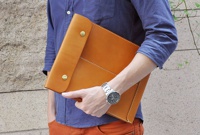 職人手作皮革信封式雙釦手拿袋，採用光滑的棕褐色皮革製成，配有兩個金色釦子和白色縫線細節，由一位穿著藍色襯衫和橘色褲子的人士持握。