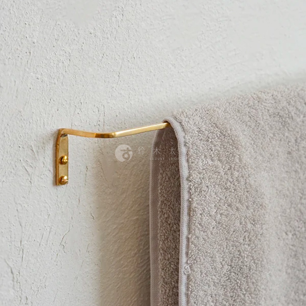 黃銅材質單桿毛巾架，L型彎曲設計，固定於白色紋理牆面，掛有淺灰色毛巾。