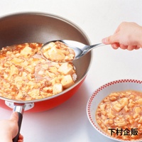 一只手正在用不鏽鋼料理分食匙從紅色的鍋中舀起麻婆豆腐，另一隻手抓住鍋柄。旁邊有一個裝有麻婆豆腐的碗。