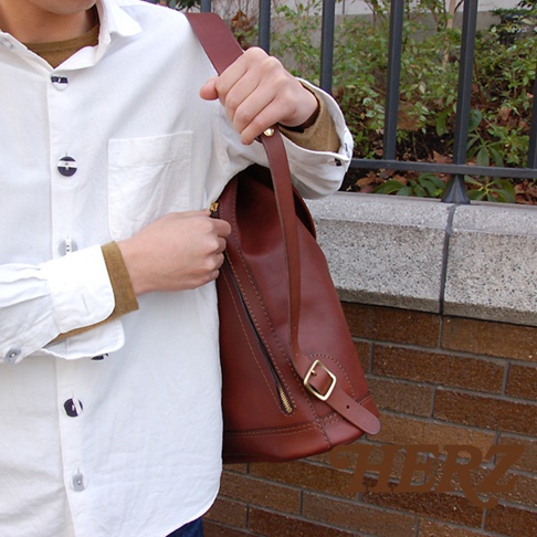 職人手作皮革單扣束口後背包，採用深棕色皮革製作，設有側邊拉鍊及可調節式單肩背帶，搭配金色金屬扣環。