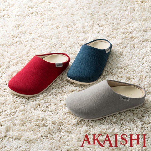 三雙減壓包覆室內拖鞋，分別為紅色、藍色和灰色，皆為無後跟設計，材質看起來是織物，放置於白色毛絨地毯上。