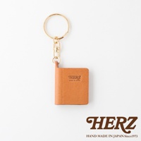 職人手作皮革書本造型鑰匙圈，採用天然皮革材質，呈現溫潤的焦糖棕色。金屬扣環和鏈條設計，方便吊掛。書本封面壓印有「HERZ」品牌標誌及「MADE IN JAPAN」字樣。