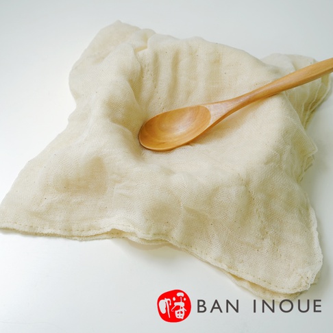 一塊米白色、織紋明顯的有機蚊帳棉廚房擦拭巾，尺寸約為 58x58 公分，上面放置著一支木製湯匙。