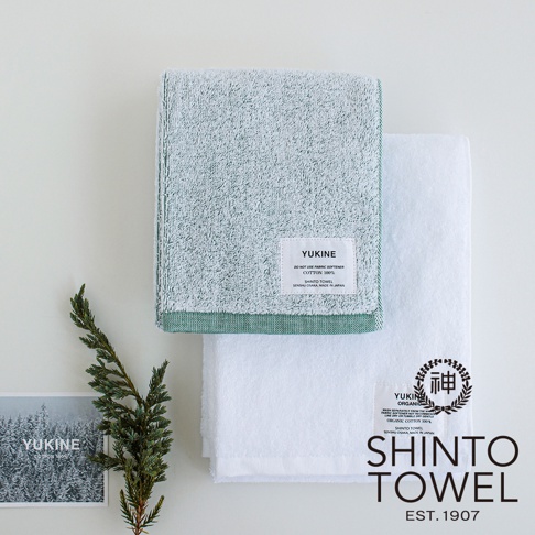 兩條 YUKINE 雪中漫步有機棉蓬厚迷你浴巾，一條為淺綠色，另一條為白色。兩條浴巾均為純棉材質，帶有品牌標籤，並配有綠色植物裝飾。