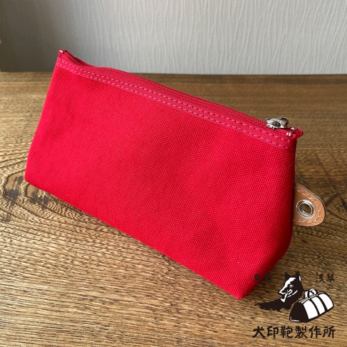紅色石洗帆布製成的小型收納包，帶有銀色金屬拉鍊，一側有一個圓形金屬孔的皮革標籤。