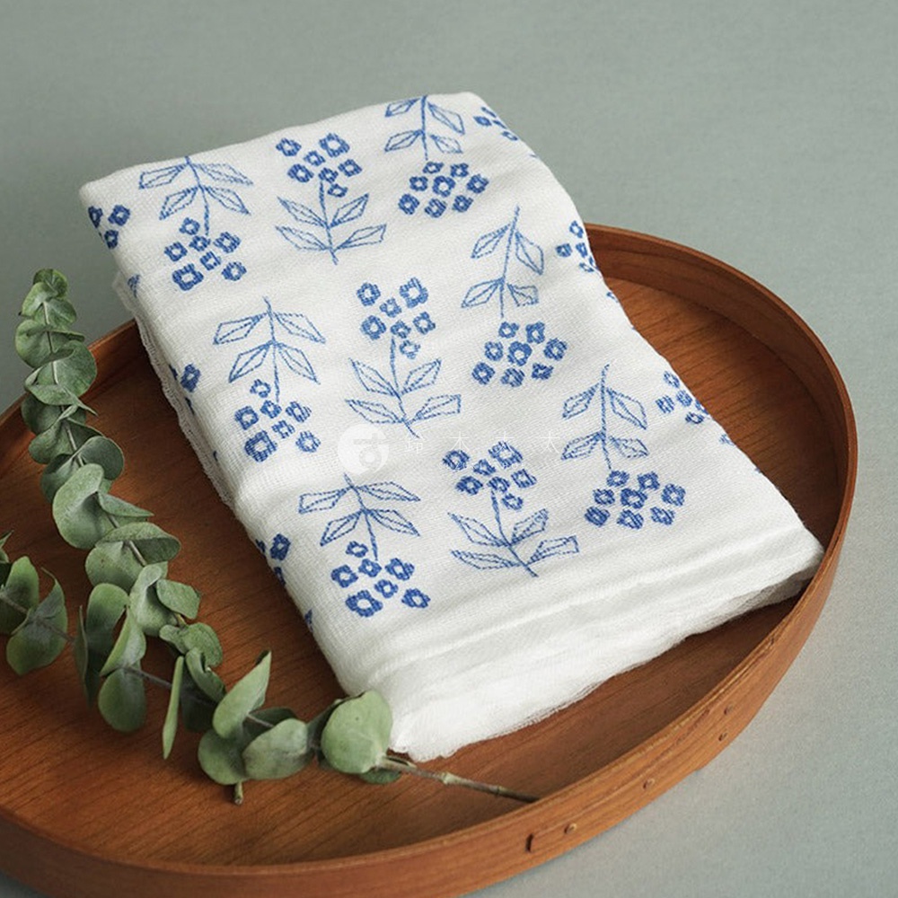 一條白色純棉花草紗布大拭巾，印有藍色小花圖案，置於木質托盤上，旁有綠葉裝飾。